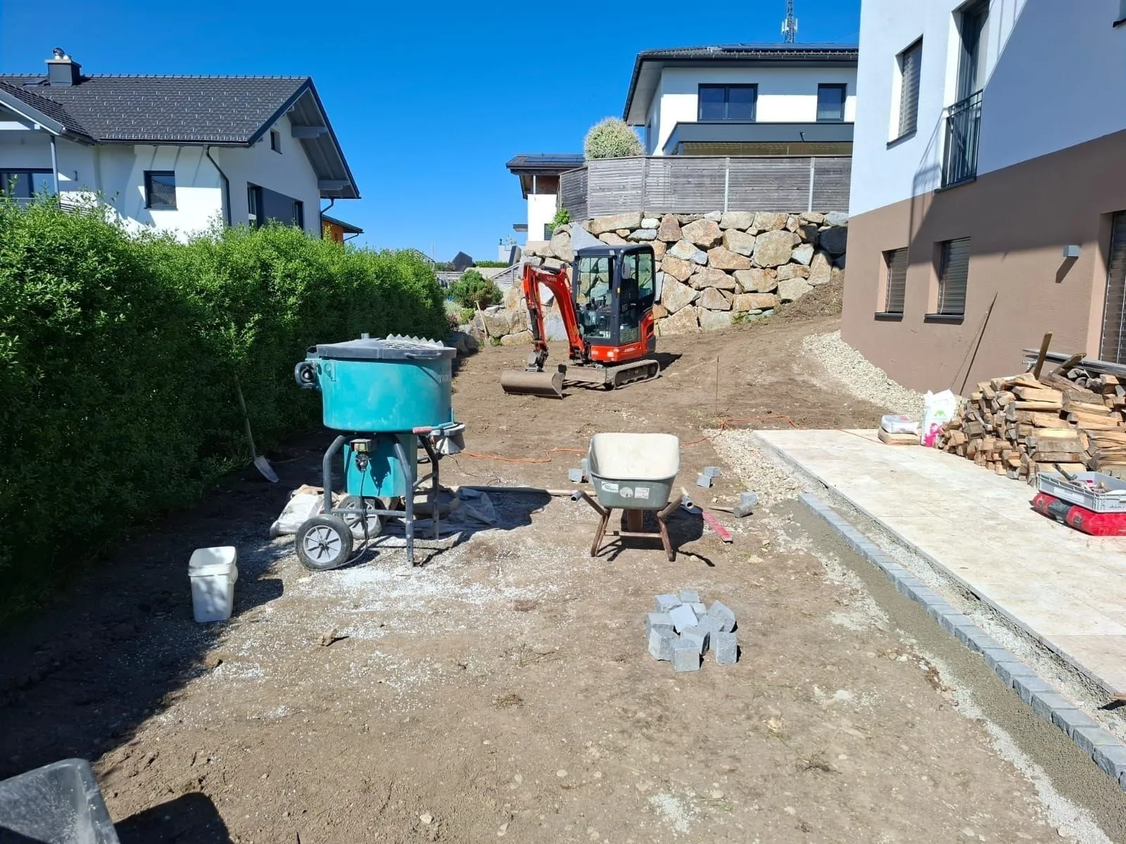 Minibagger auf einer Gartenbaustelle neben Mischer, Schubkarre und frisch verlegtem Plattenbelag
