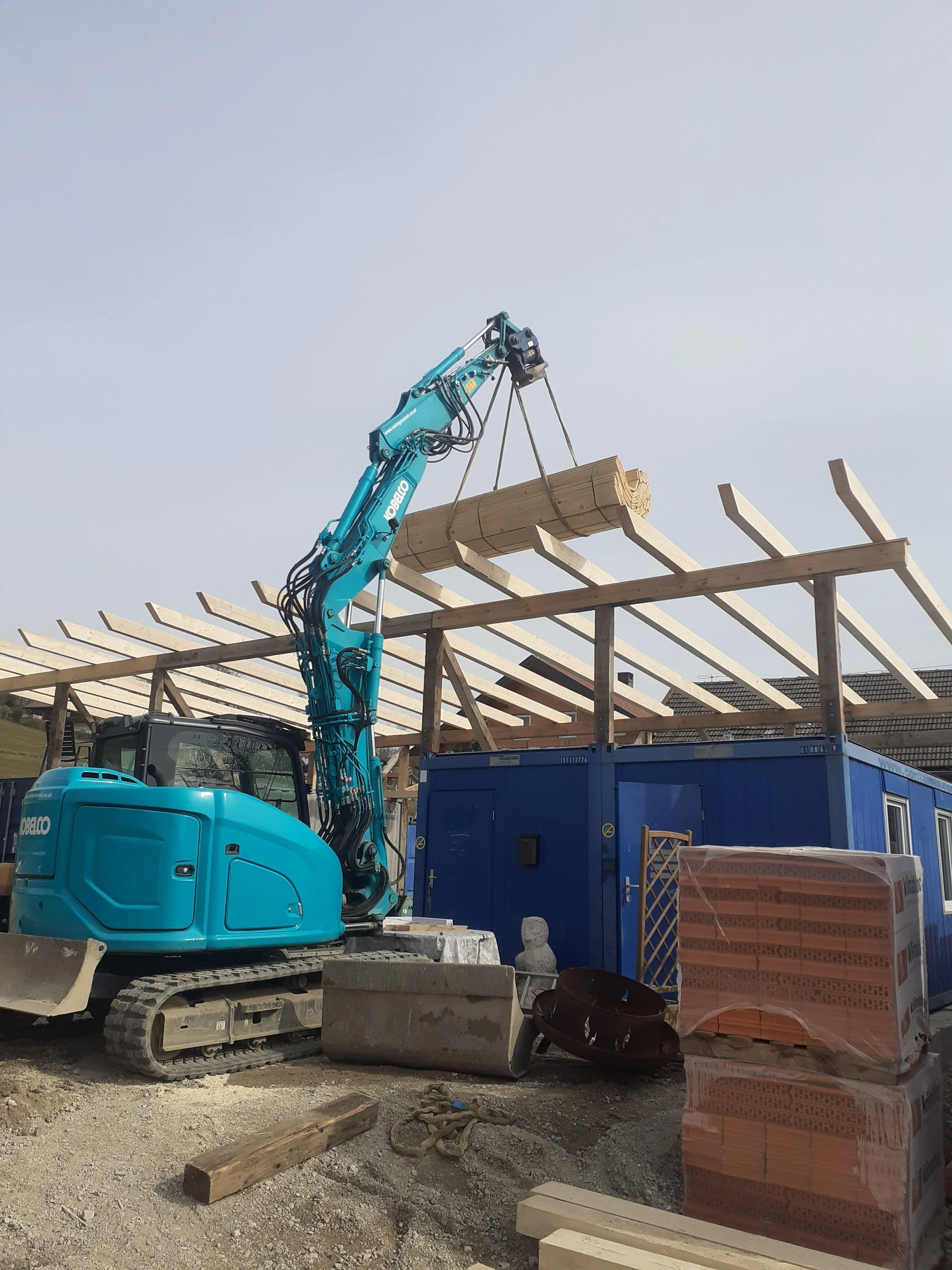 Kettenbagger auf einer Baustelle neben Baucontainern und Dachstuhlholz