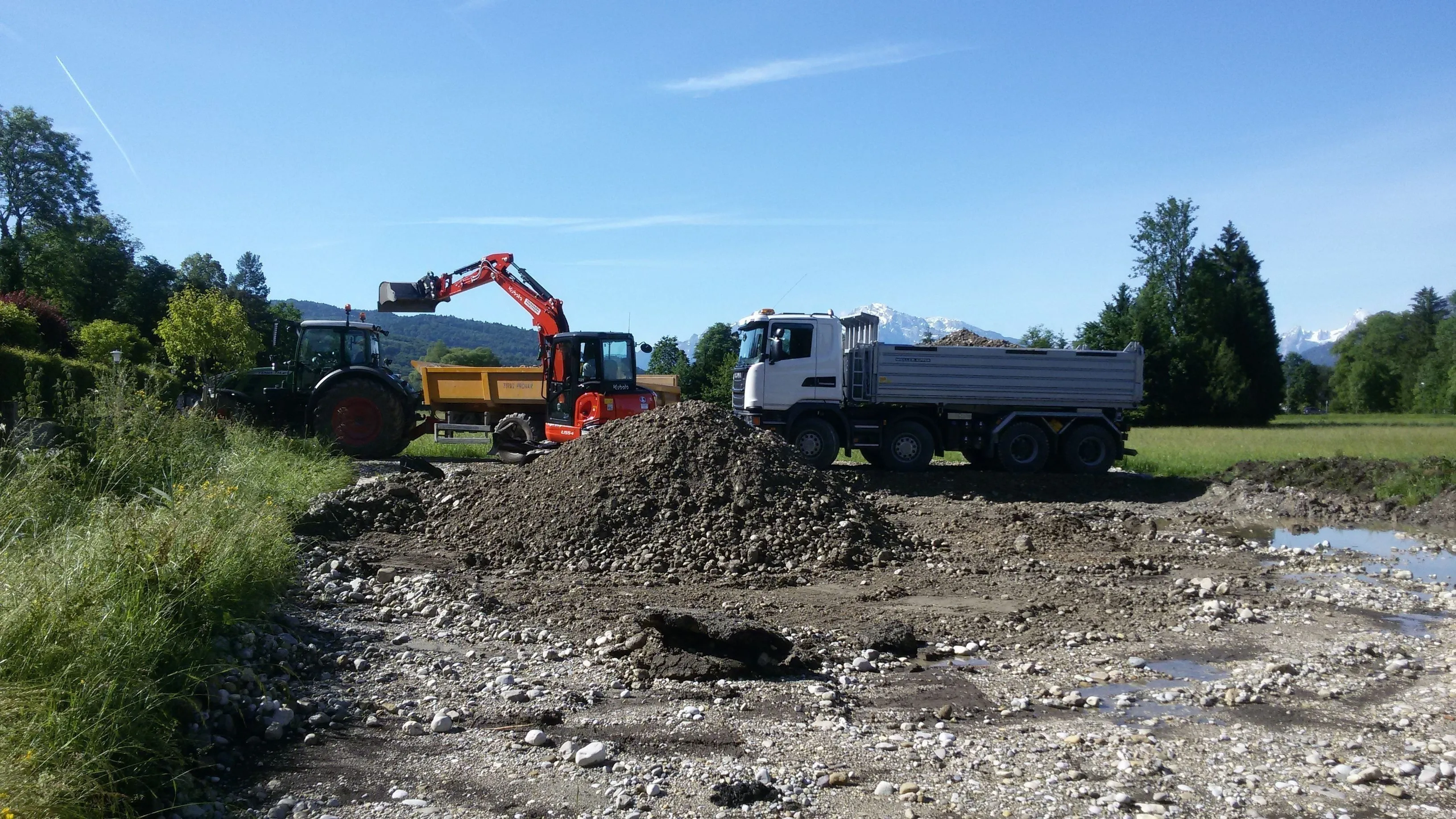 Bagger belädt Traktor-Kipper, daneben wartet ein Kipper-LKW