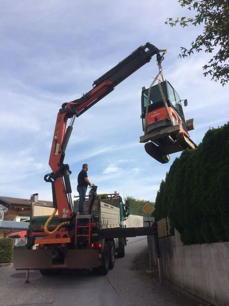 Ladekran hebt einen Minibagger über eine hohe Hecke