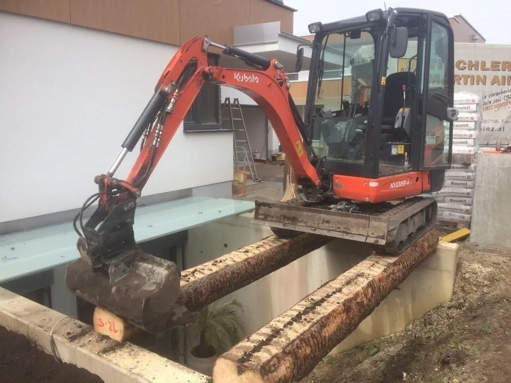 Minibagger arbeitet auf Baumstämmen stehend über einer betonierten Öffnung