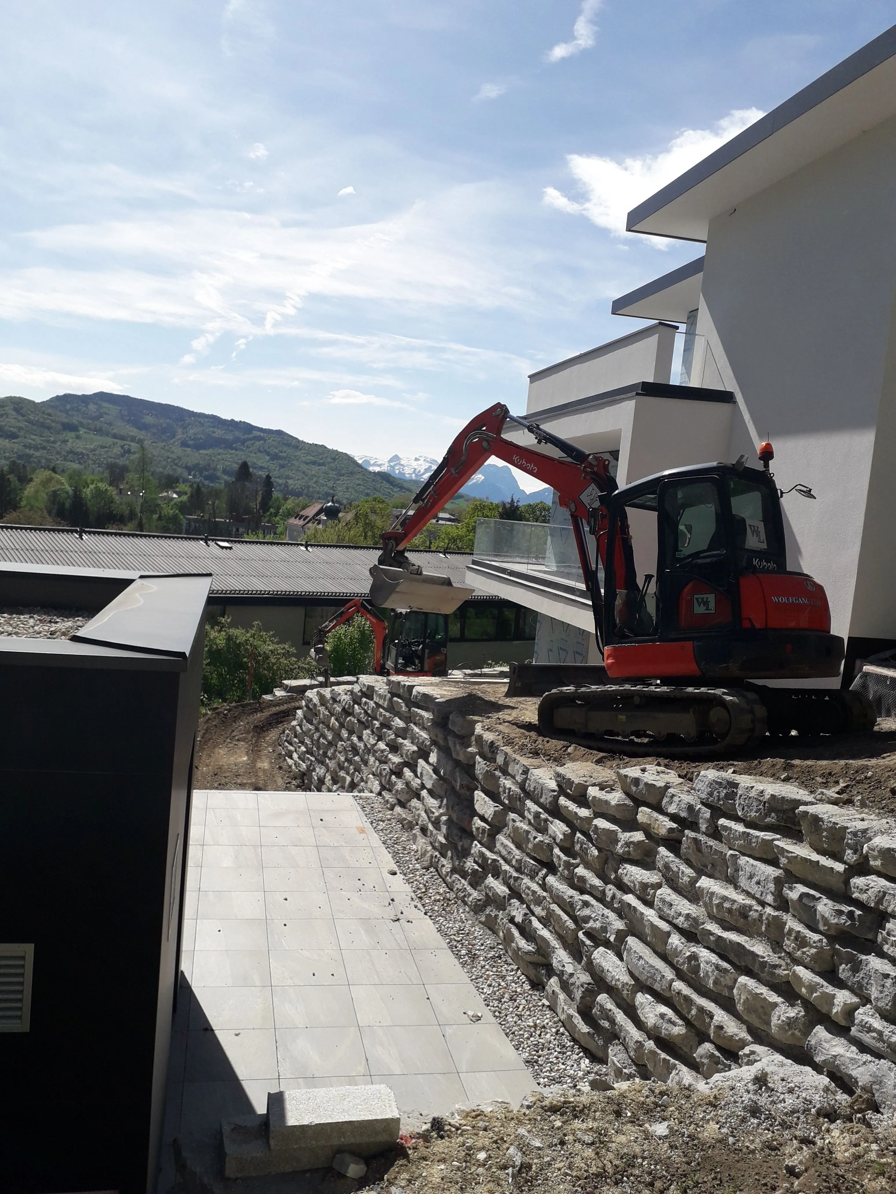 Minibagger bei Arbeiten an einer geschlichteten Natursteinmauer an einem modernen Neubau