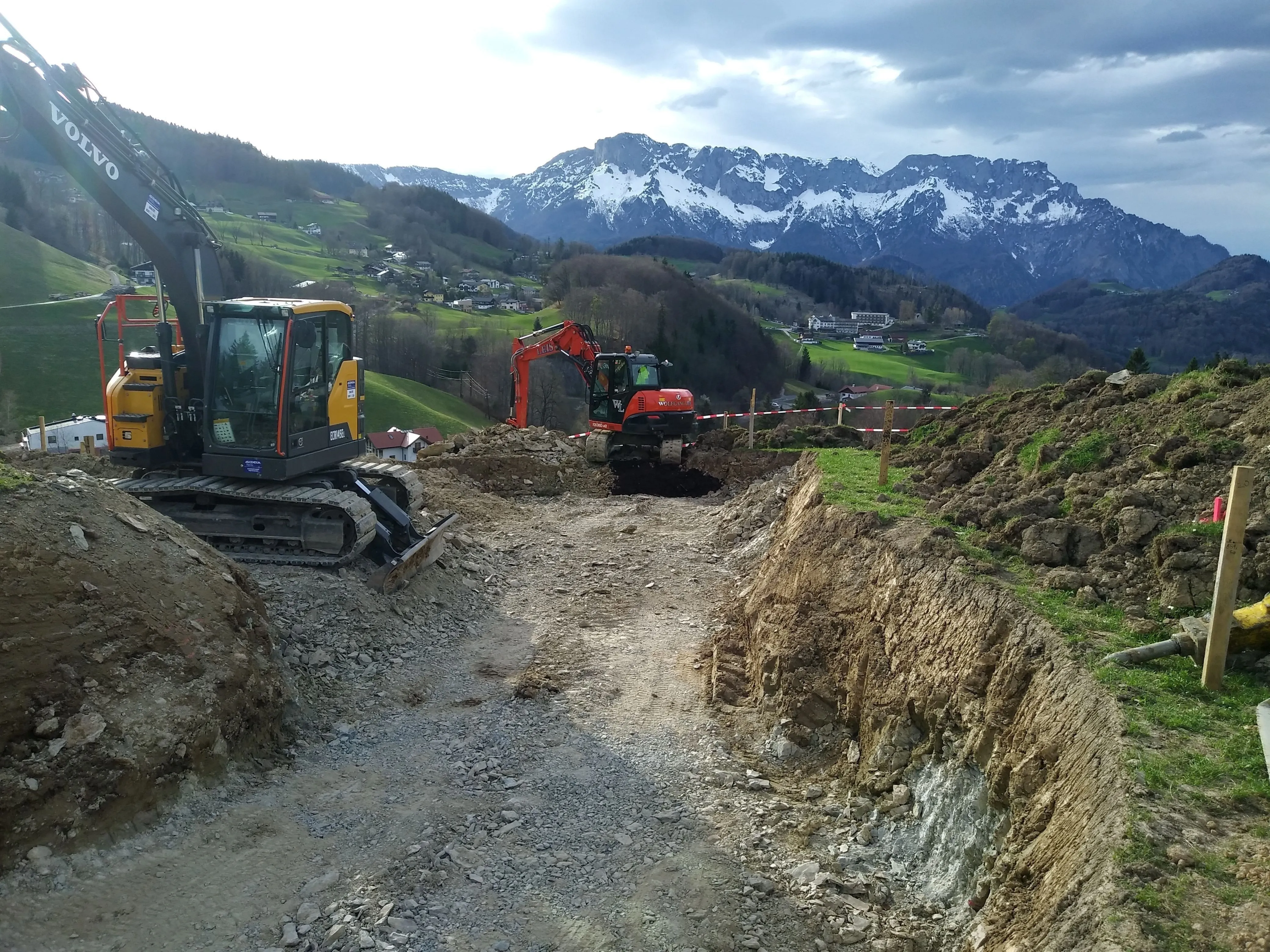 Zwei Bagger bauen einen Weg am Hang, dahinter Alpenpanorama