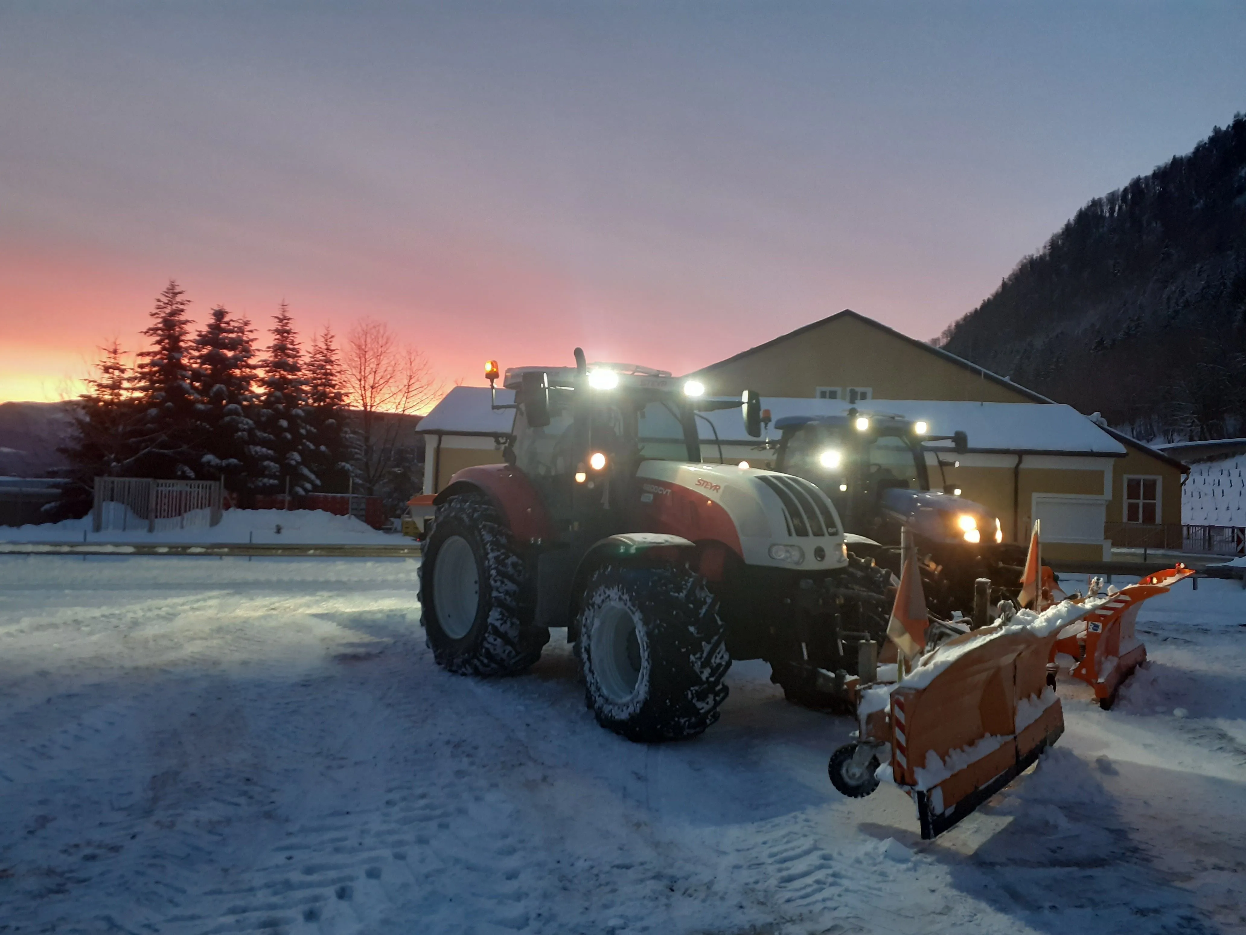 Traktor mit Schneepflug und Streugerät auf schneebedecktem Platz im Morgengrauen